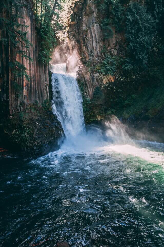 Ein Fluss und ein Wasserfall im Wald. Einige Sonnenstrahlen außerdem im Hintergrund. 