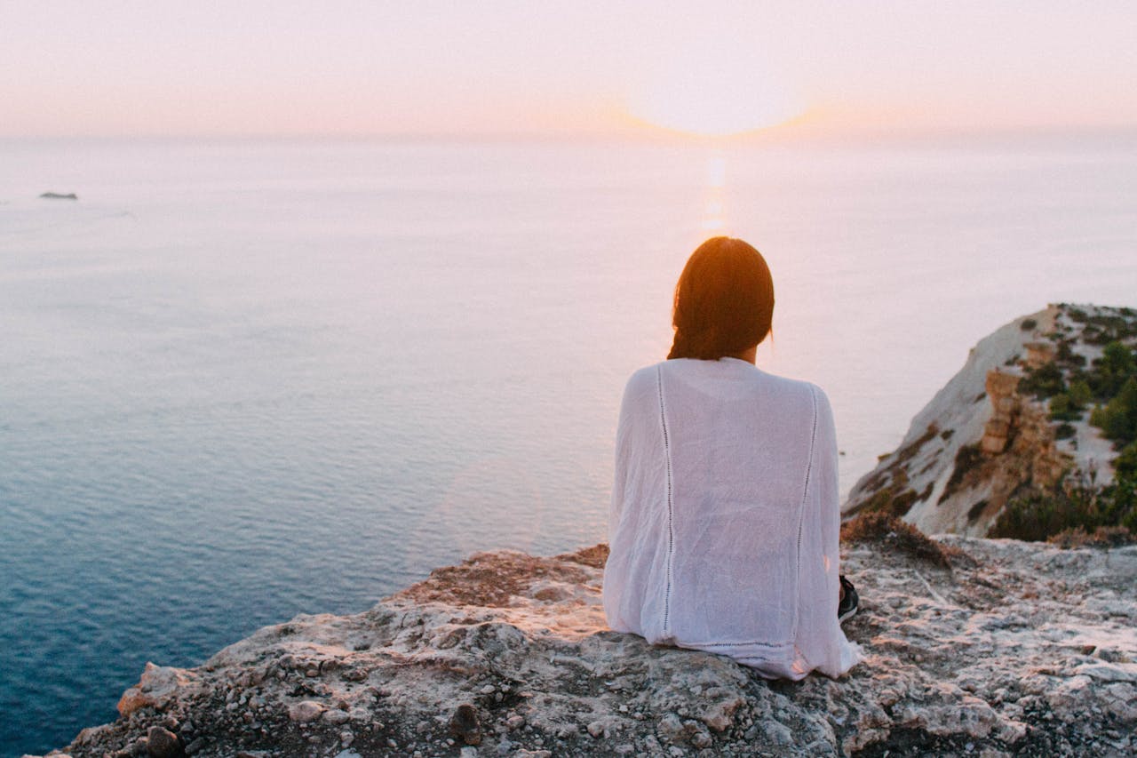 Achtsam leben, im Einklang mit der Natur - ungefähr das vermittelt die Silhouette einer Frau in der Abendsonne, sie sitzt auf einem Felsen und blickt auf das Meer.