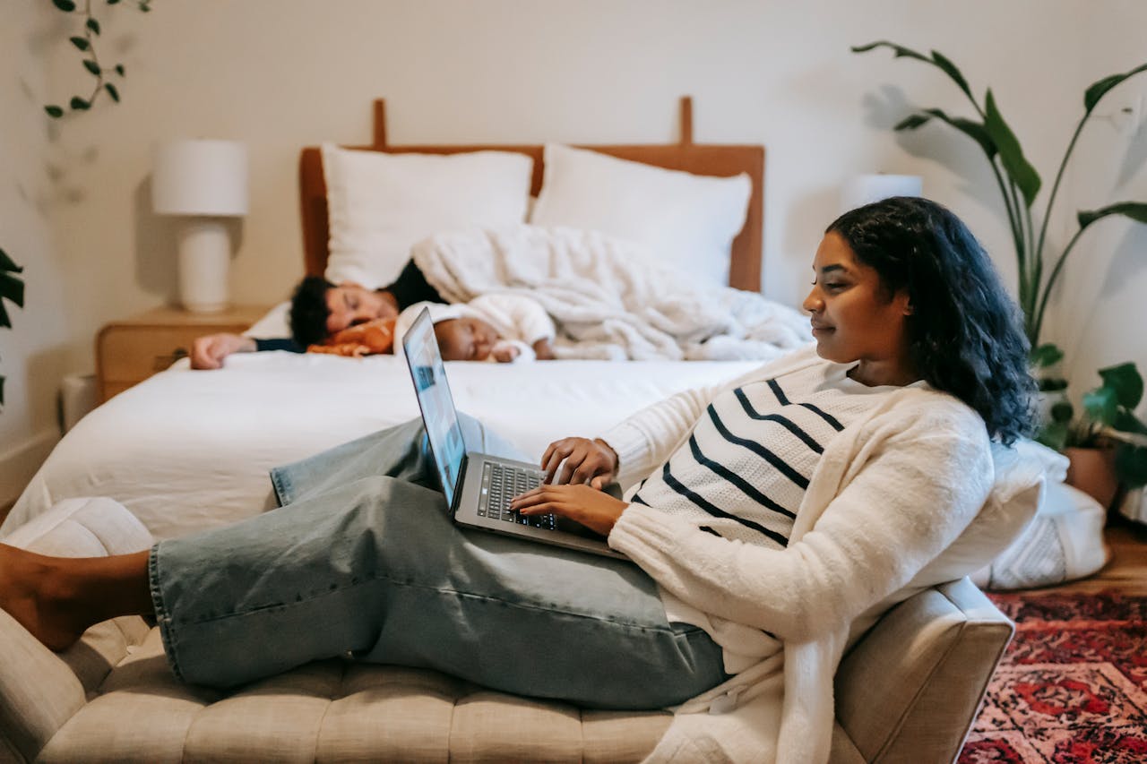 Ein Schlafzimmer, auf dem Bett liegen Mann und Kind und schlafen. Die Frau sitzt davor und arbeitet an ihrem Laptop. So kann Care Arbeit und Familie geteilt werden.