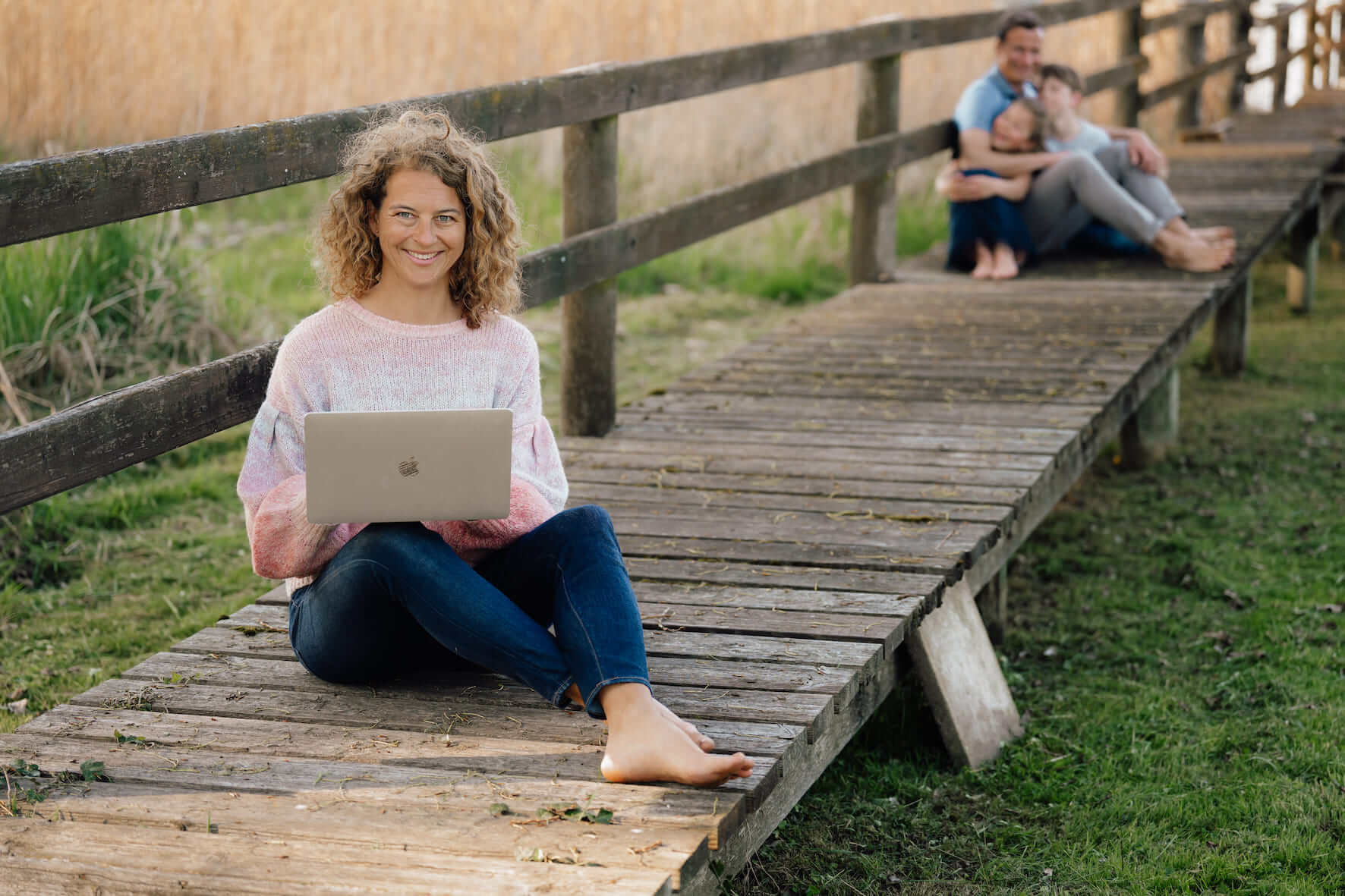 Coaching Katrin Fuchs sitzt auf einem Steg in der Natur. Im verschwommenen Hintergrund sind ihr Mann und Kinder zu sehen. 