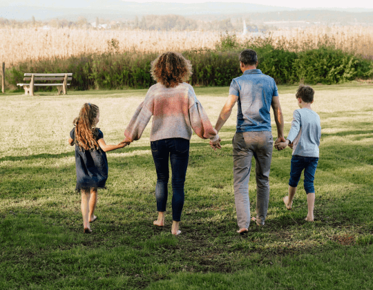 Katrin Fuchs mit Mann und ihren beiden Kindern. Sie sind von hinten zu sehen, halten sich an den Händen und laufen barfuß über eine Wiese. Sie zeigen, wie Care Arbeit und Familie gleichberechtigt gehen kann.