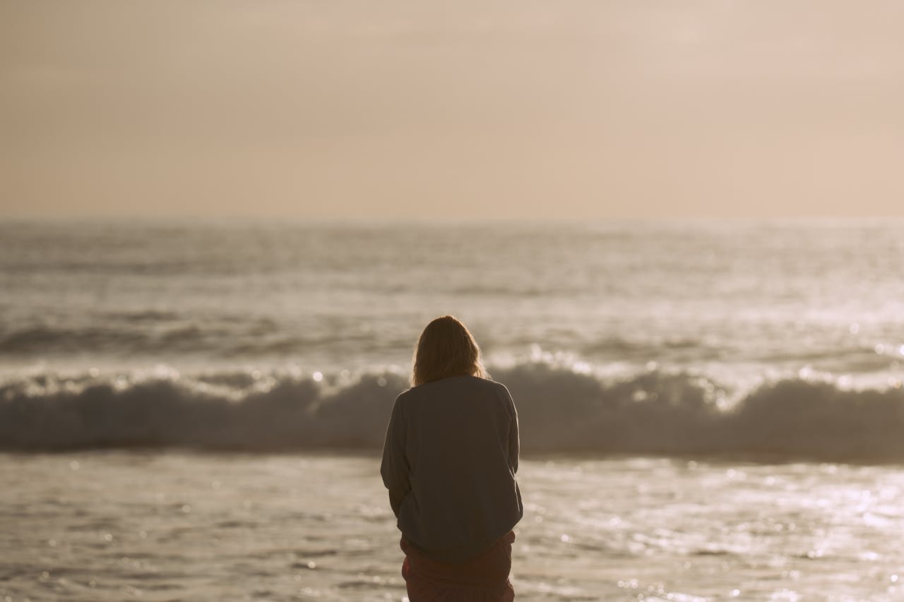 Eine Frau steht in der Abendsonne am Meer. Sie sinniert vielleicht über Sprüche Achtsamkeit. 