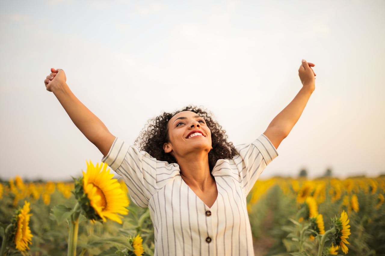 Eine Frau steht in einem Sonnenblumen-Feld und jubelt. Sie zeigt ihr authentisches Ich auf ihrer Über mich Seite. 